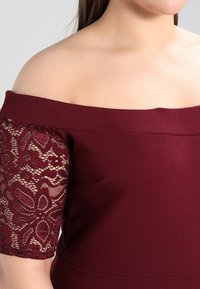 Bordeauxrotes Off-Shoulder-Oberteil mit Spitzenärmel-Detail, aus strukturiertem Stoff und tailliertem Schnitt, der offene Schultern betont.