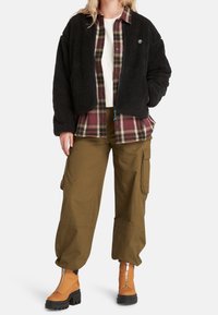 Czarna kurtka z polaru na koszulę w burgundowo-beżową kratę, zestawiona z oliwkowymi spodniami typu cargo i pomarańczowymi botkami z czarnymi akcentami.