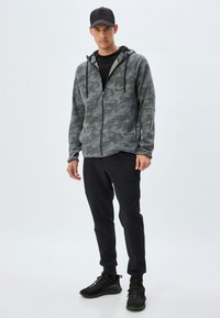 Sudadera con capucha de camuflaje gris con acentos negros, combinada con joggers negros ajustados y zapatillas deportivas negras. Tejido texturizado, corte casual.