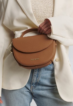Sac en cuir marron à texture pebble avec un design en forme de rabat incurvé et un logo doré. Il dispose d'une petite poignée et est posé contre un blazer blanc.
