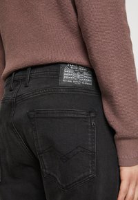 Črne denim hlače z teksturiranim pasom, ki vsebuje oznako z arhitekturnim dizajnom. Zadnji žepi imajo izrazite šive.