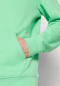 Felpa verde menta realizzata in tessuto morbido, con tasca anteriore e polsini a coste. Cuciture decorative visibili lungo i bordi.