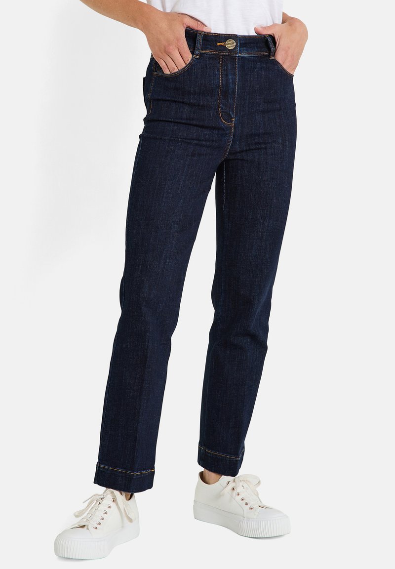 Jeans de mezclilla azul oscuro con corte recto, con costuras naranjas, bolsillos frontales y cierre de botón. Combinados con zapatillas blancas.