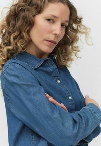 Koszula dżinsowa w średnim niebieskim odcieniu, z kołnierzykiem, zapinana na guziki, z długim rękawem. Tkanina ma gładką fakturę z delikatnym wyblaknięciem.