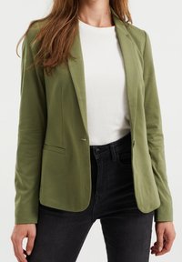 Olivgrön kavaj med skräddarsydd passform, slagkragar, en knapp och framfickor. Buren över en vit skjorta och svarta jeans.