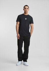 Zwarte katoenen T-shirt met een rond logo op de borst, gecombineerd met zwarte cargobroeken en witte sneakers. Minimalistische stijl, zonder patronen.