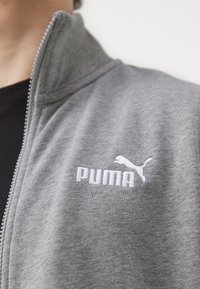Veste zippée grise avec un col haut, fabriquée en tissu doux. Présente un logo "PUMA" brodé en blanc et un design de chat sautant.