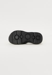 Svart gummi sneaker såle med teksturert grep, sirkulære og rektangulære mønstre, og hevede aksenter for bedre feste. Flat, svakt avrundet form.