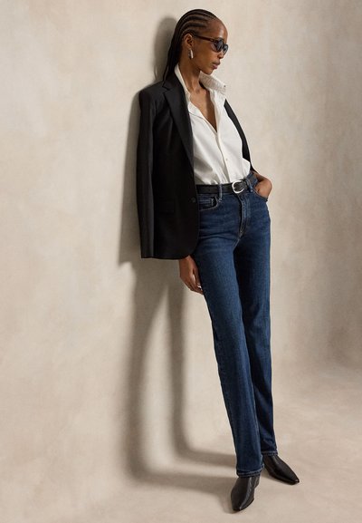Donna con occhiali da sole, camicia bianca, blazer nero sulle spalle, jeans blu scuro e stivali neri a punta, appoggiata a un muro beige.