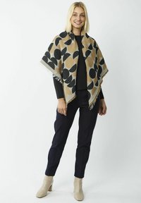 Geweven sjaal in bruin met grote zwarte en lichtbruine cirkelpatronen, met franjes aan de randen, gedragen over een donkere top en gecombineerd met donkere broek en beige laarzen.