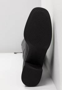 Černá kožená bota zobrazená z paty, s texturovanou gumovou podrážkou s horizontálními drážkami a mírným podpatkem.