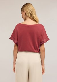 Top de manga corta en color burdeos con un corte holgado y acentos plisados en las mangas. Combinado con pantalones beige de pierna ancha.