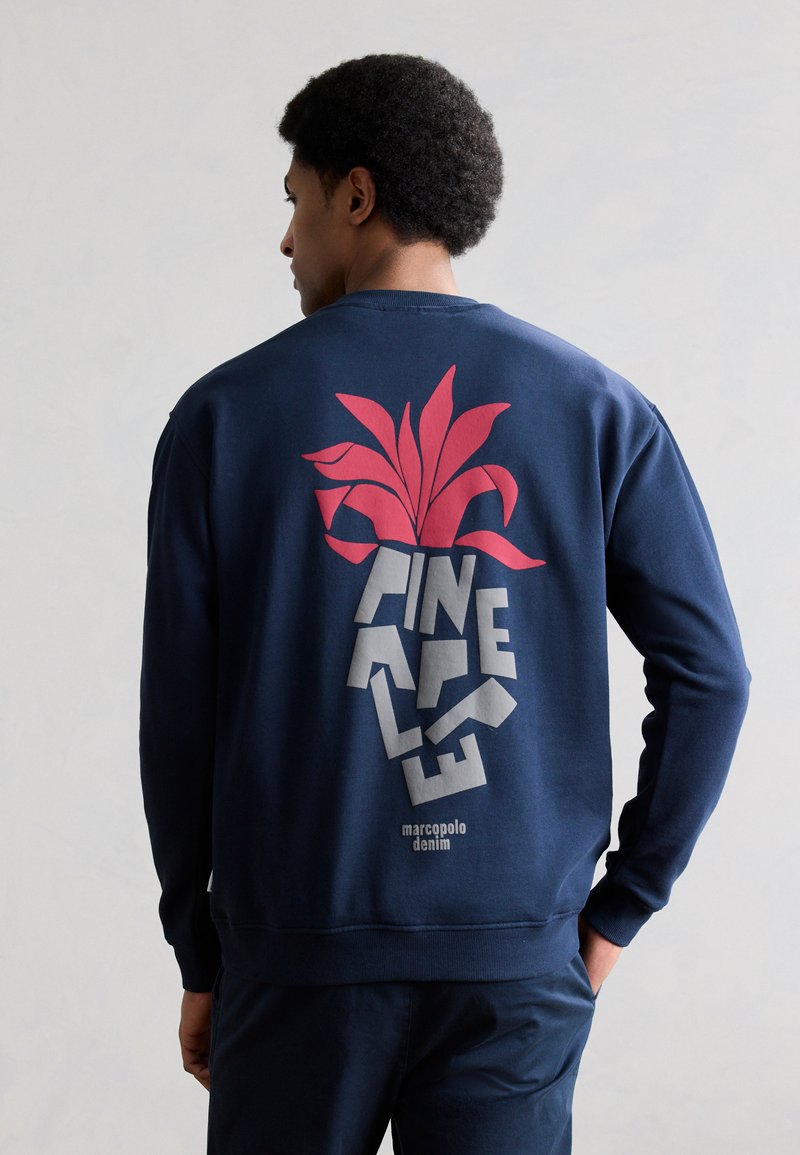 Άνδρας που φοράει ναυτικό φούτερ με κόκκινο γραφικό ανανά και κείμενο «PINAPPLE» στην πλάτη, στέκεται με το χέρι στην τσέπη, κοιτάζοντας προς τα αριστερά.