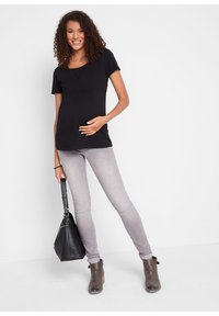 Maglietta nera per maternità con maniche corte, arricciata sotto il busto. Abbinata a jeans skinny grigi chiari e stivali alla caviglia grigi. Tiene in mano una borsa nera.