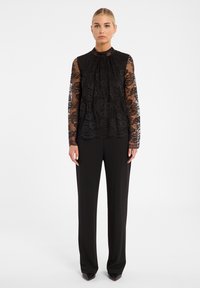 Zwarte kanten blouse met lange mouwen en een hoge hals, voorzien van bloemenpatronen en plooien, gecombineerd met zwarte wijde broek.