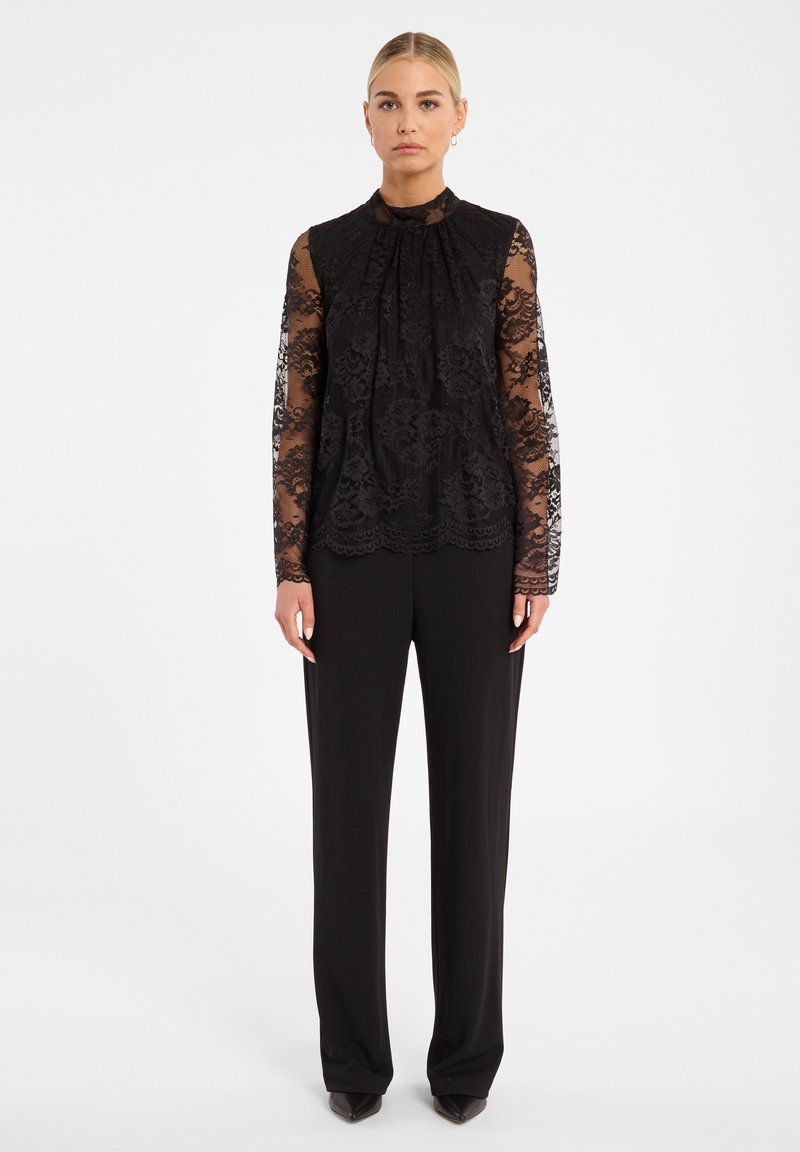 Zwarte kanten blouse met lange mouwen en een hoge hals, voorzien van bloemenpatronen en plooien, gecombineerd met zwarte wijde broek.