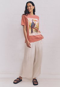 Smiling woman stands wearing a brick-red T-shirt with a toucan graphic, loose beige pants, black buckle sandals, and a gold bracelet.