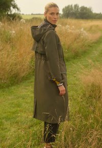 olivově zelená voděodolná pláštěnka s kapucí s leopardím vzorem, stahovaným pasem a postranními kapsami. Vyznačuje se dlouhým, rovně střiženým designem a jemnými akcenty.