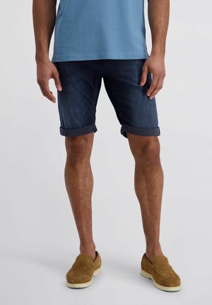 Man die donker denim shorts met opgerolde pijpen, een blauw overhemd en beige suède instapschoenen draagt, staand tegen een effen achtergrond.