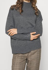 Frau in einem grauen gerippten Strickpullover mit hohem Kragen und karierten Hosen, die die Arme an der Taille verschränkt.