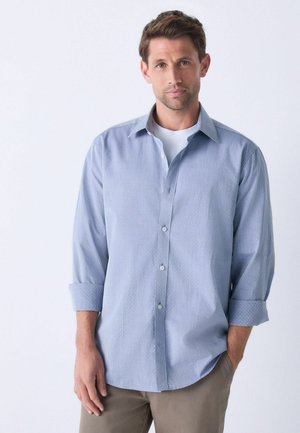 Helles blaues Button-Down-Hemd mit einem dezenten gepunkteten Muster, langärmlig und an den Manschetten aufgerollt, kombiniert mit einem weißen Rundhals-T-Shirt darunter.
