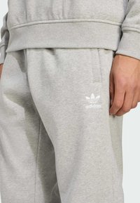 Svetlo šedé tepláky Adidas s bočnými vreckami, s bielym logom na ľavom stehne a mäkkou, textúrovanou tkaninou.