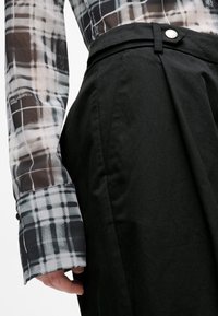 Langärmliges Hemd mit einem schwarz-weißen Karomuster, aus durchscheinendem Stoff gefertigt. Kombiniert mit hoch taillierten schwarzen Hosen mit Knopfverschluss.