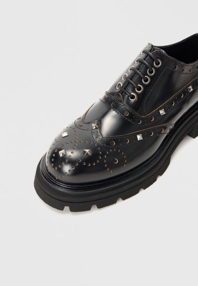 Pantofii brogue din piele neagră, cu accente cu cuie, perforații decorative, talpă plată din cauciuc negru și design cu vârf rotund, prevăzuți cu șireturi.