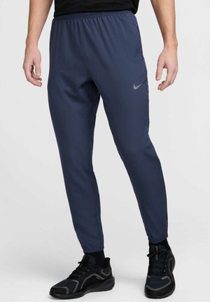 Granatowe sportowe spodnie z elastycznym pasem i ściągaczami przy kostkach. Na lewej udzie znajduje się szare logo Nike. Gładka faktura materiału.