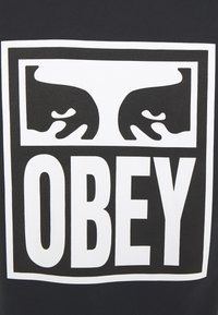 Černá látka s velkým bílým obdélníkovým grafickým motivem, na kterém jsou stylizované ruce nad slovem „OBEY“ napsaným tučnými černými velkými písmeny.