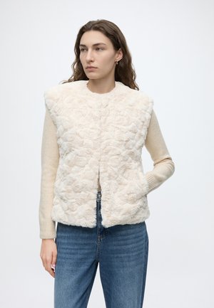 Femme aux longs cheveux bruns porte une veste en fausse fourrure crème texturée par-dessus un pull beige et un jean bleu, debout avec une main dans la poche de la veste.