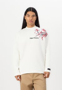 Sweatshirt blanc à manches longues avec broderie florale rose sur l'épaule gauche et texte "CARLO COLUCCI". Tissu lisse, coupe décontractée.