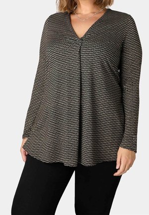 Mujer con camiseta de manga larga blanca y negra con estampado en V con detalle de pliegues combinada con pantalones negros lisos sobre fondo claro.