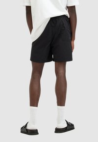 Black swim shorts made of lightweight fabric with an elastic waistband and side pocket, paired with black slide sandals and white crew socks.
