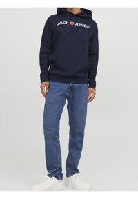 Námořnicky modrá mikina s kapucí s nápisem "JACK & JONES", klokaní kapsou a nastavitelným stahovacím šňůrkou, doplněná světle modrými džínami a bílými teniskami.