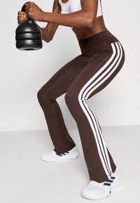 Pantalon marron évasé avec trois bandes blanches verticales sur chaque jambe. Le matériau semble lisse et extensible. Une kettlebell noire est tenue dans la main gauche.
