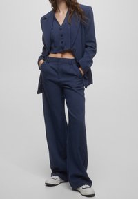 Marynarski krótki żakiet i dopasowane wysokie szerokie spodnie, oba w gładkim materiale z wyraźnym krojem i wieloma guzikami.