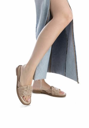 Sandalias deslizables trenzadas en color tostado con suela plana, que presentan un diseño de tiras cruzadas hechas de material texturizado. Falda de mezclilla parcialmente visible con aberturas laterales.