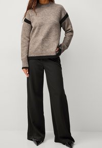 Pull en tricot brun avec des rayures noires aux épaules, des poignets et un ourlet côtelés. Associé à un pantalon large noir et des chaussures à bouts pointus noires.