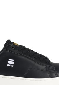 Black leather sneaker with white rubber sole, featuring flat black laces, a logo on the side, and a gold accent tag on the upper.