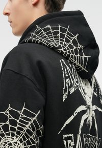 Sāniskats uz personu, kas valkā melnu kapuci ar baltām zirnekļa un tīkla grafikām aizmugurē, piedurknēs un uz kapuces.