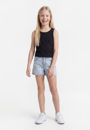 Uśmiechnięta dziewczyna z długimi blond włosami stoi, ubrana w czarny top bez rękawów, jasne krótkie spodenki jeansowe i białe trampki na białym tle.
