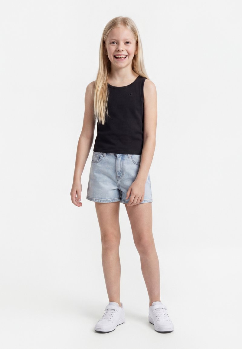 Fille souriante aux longs cheveux blonds, portant un haut noir sans manches, un short en jean clair et des baskets blanches sur un fond blanc.