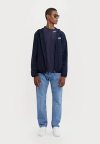 Marine vindjakke med hætte, parret med en mørk T-shirt, lyseblå jeans og hvide sneakers. Inkluderer solbriller, ingen mønstre eller detaljer.
