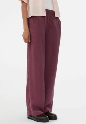 Persoon draagt losse, hoog getailleerde bordeauxrode broek met zijzakken en zwarte plateauzolen, gecombineerd met een lichtroze kort mouw shirt.