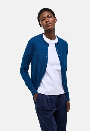 Femme portant un cardigan bleu boutonné sur une chemise blanche et un pantalon plissé bleu marine, debout avec une main dans la poche.