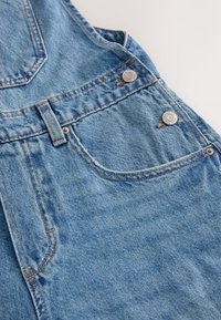Detaliu al blugilor albastre din denim, arătând buzunarul frontal, bucla pentru curea și nituri argintii pe un fundal texturat din țesătură.