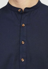 Camicia blu navy con colletto a banda, dotata di quattro bottoni in finitura legno marrone, realizzata in un tessuto liscio con un leggero lucido.