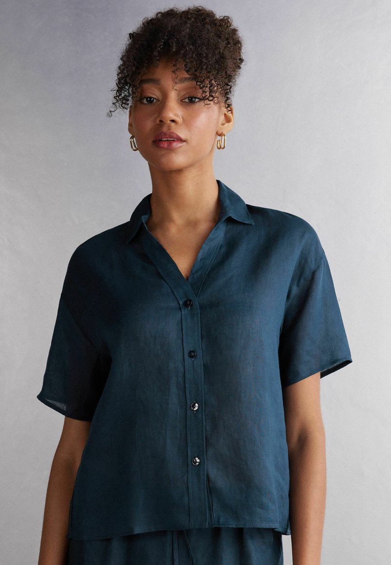 Camisa de lino azul oscuro de manga corta con cuello, cierre de botones en la parte delantera y un corte relajado, que presenta un ligero brillo y una textura sutil.