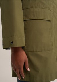 Olive green parka with a smooth texture, featuring a side pocket and snap fasteners at the cuff. Close-up of sleeve and hand.